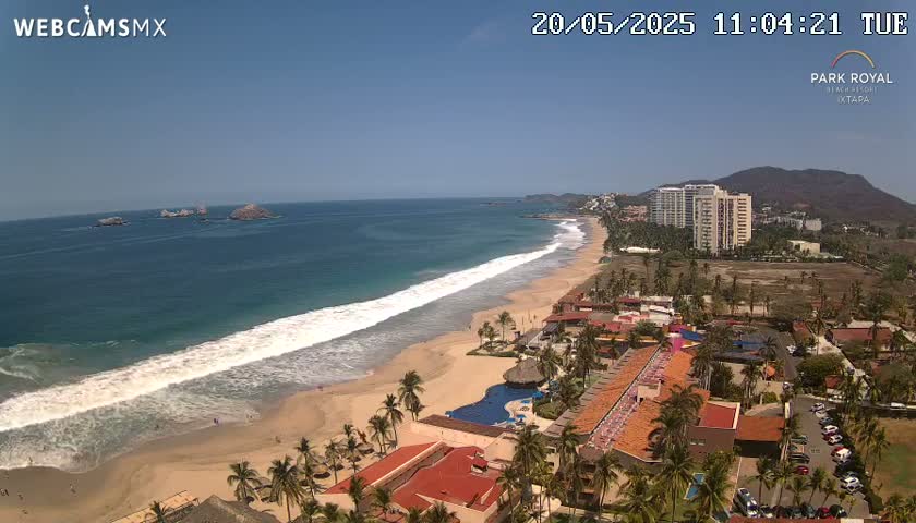 An aerial view shows a long sandy beach with waves lapping the shore, a resort with red-tiled roofs, and high-rise buildings in the background under a clear, sunny sky.