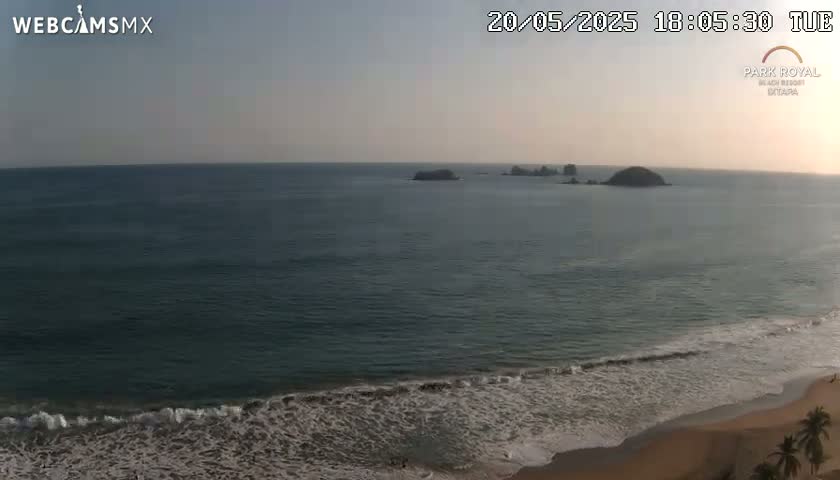 A calm ocean with small waves laps a sandy beach under a clear, dusky sky, with a few small islands visible in the distance.
