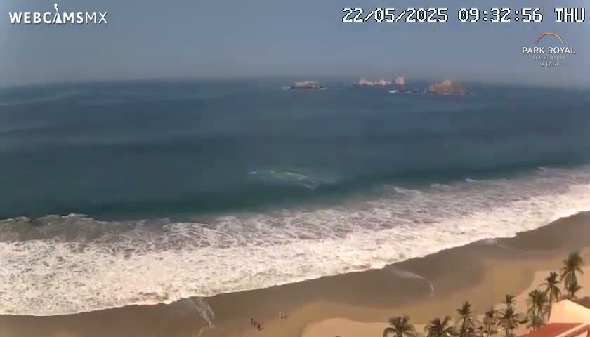 A sandy beach with waves rolling in under a mostly clear sky, and a few small islands visible in the distance.