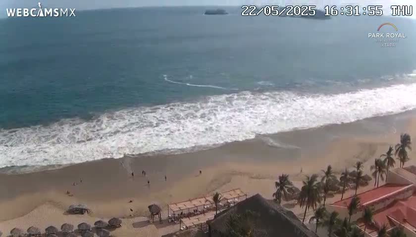 An aerial view shows a sandy beach with moderate waves crashing on the shore, palm trees, and a resort building under an overcast sky.