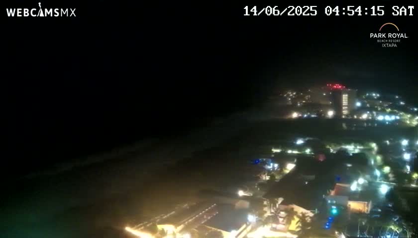 A nighttime, high-angle view shows a coastal town illuminated with lights, extending to a dark ocean under a cloudy sky.