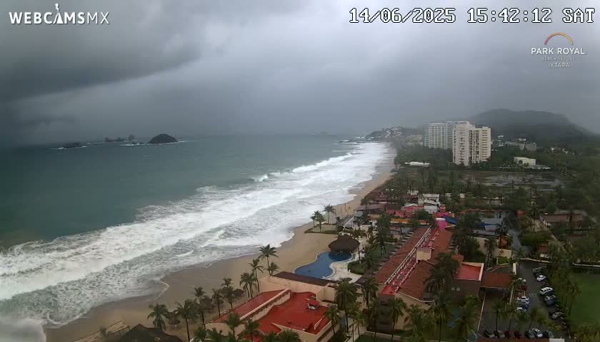 Large waves crash on a sandy beach near a resort with palm trees under stormy conditions.