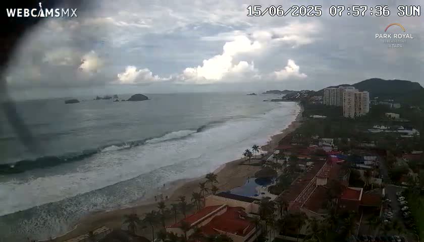 An ocean beach scene under a cloudy sky shows waves breaking on a sandy shore near hotels and buildings, with islands visible in the distance.