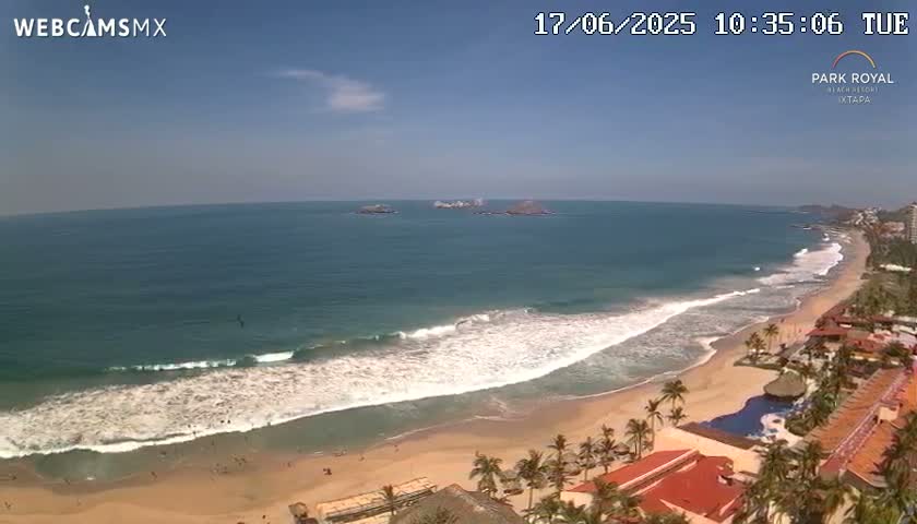 An aerial view shows a sandy beach with gentle waves lapping the shore under a mostly sunny sky, with a resort visible along the coastline and small islands in the distance.
