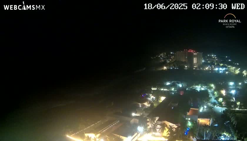 A nighttime, aerial view of a resort town shows a dark ocean, illuminated buildings, and palm trees under a clear, dark sky.