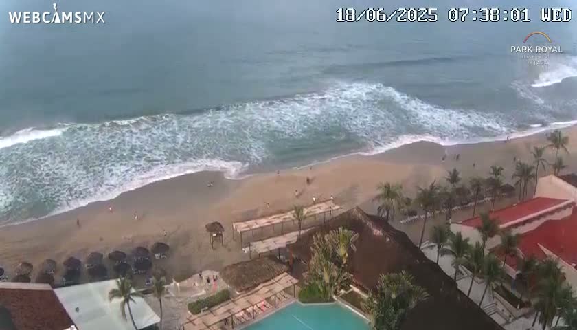 A beach with moderate waves under an overcast sky is seen from above, showing a resort with a swimming pool adjacent to the sandy shore.