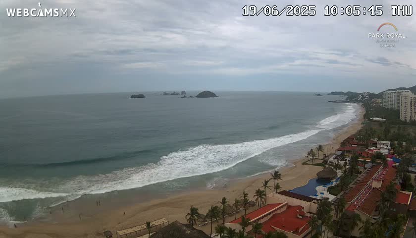 A beach with moderate waves under a cloudy sky, showing a resort area and distant islands.