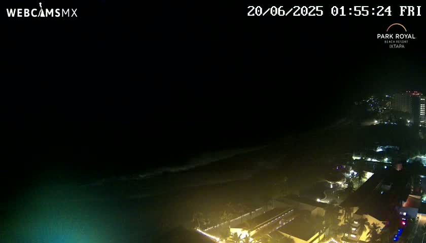 A nighttime, coastal view shows dark ocean waves lapping a beach lined with illuminated buildings.