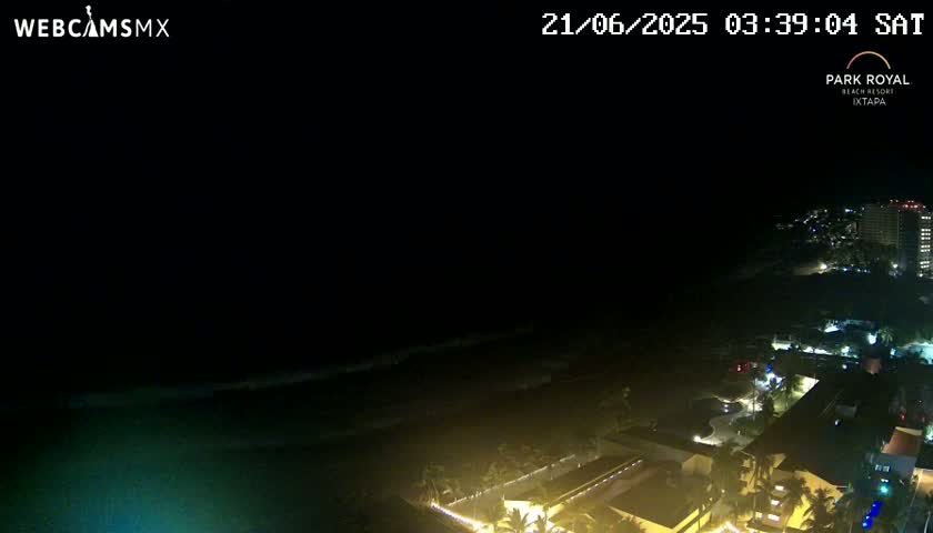 A nighttime coastal scene shows dark ocean waves lapping a shore lined with dimly lit buildings under a clear, dark sky.