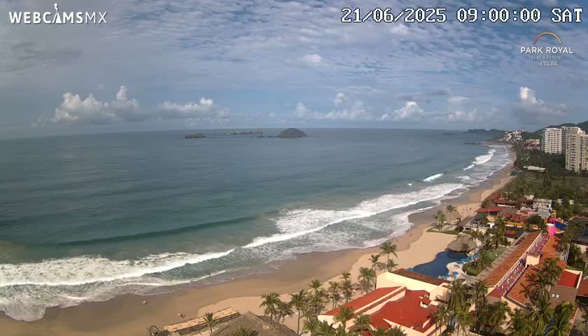 An aerial view shows a sandy beach with gentle waves lapping the shore under a partly cloudy sky, with resort buildings and palm trees lining the beach.