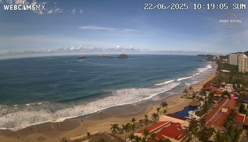An aerial view shows a sandy beach with gentle waves lapping the shore under a mostly sunny sky, with resort buildings and palm trees lining the beach.