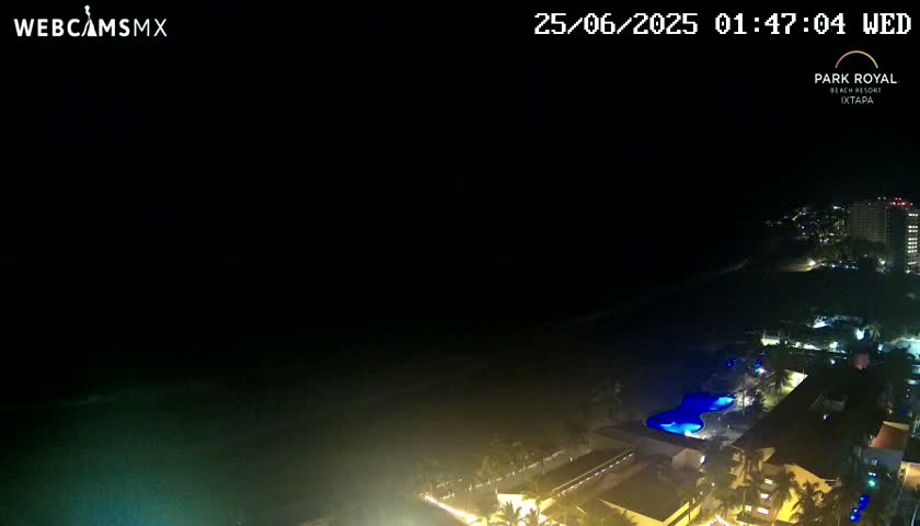 A nighttime, coastal view shows a dark ocean, a beach, and a resort complex with illuminated buildings and pools.
