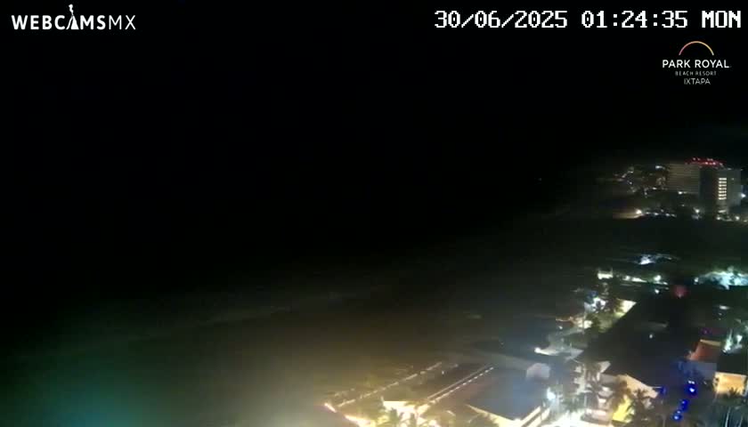 A nighttime, aerial view of a coastal city with illuminated buildings and a dark ocean under a clear sky.