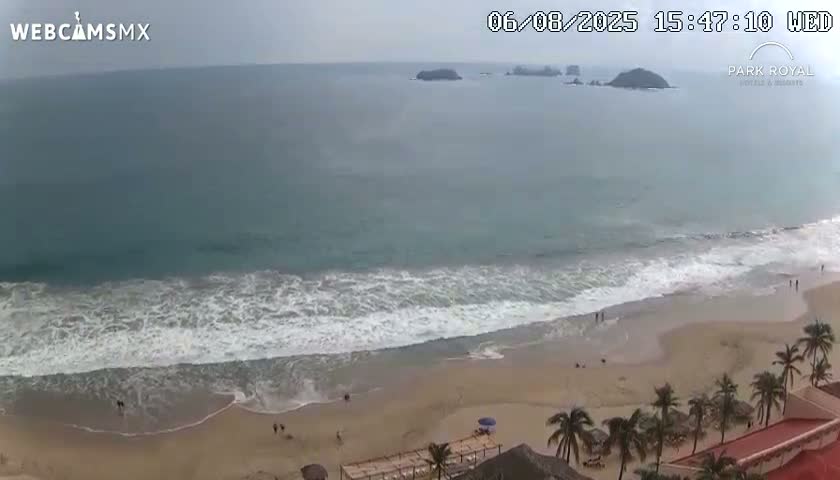 An overcast day shows a beach with moderate waves lapping the shore, a few people walking on the sand, and palm trees and buildings in the foreground, with islands visible in the distance.