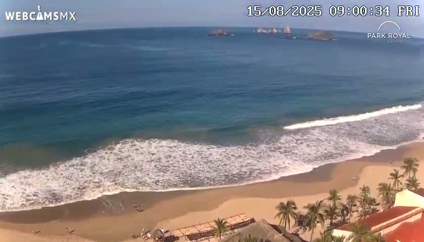 An overcast day shows a beach with moderate waves lapping the shore, a few people walking on the sand, and palm trees and buildings in the foreground, with islands visible in the distance.