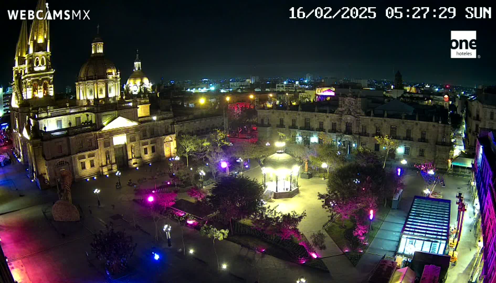 A brightly lit plaza at night, featuring a large cathedral, surrounding buildings, a gazebo, and colorful lights illuminating the scene.