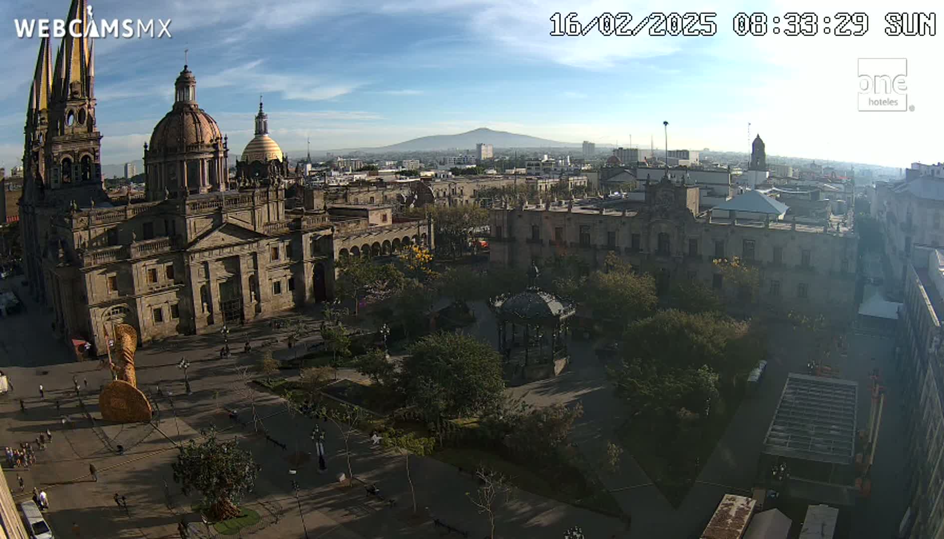 A large cathedral and surrounding plaza are shown on a sunny day with a mountain visible in the distance.