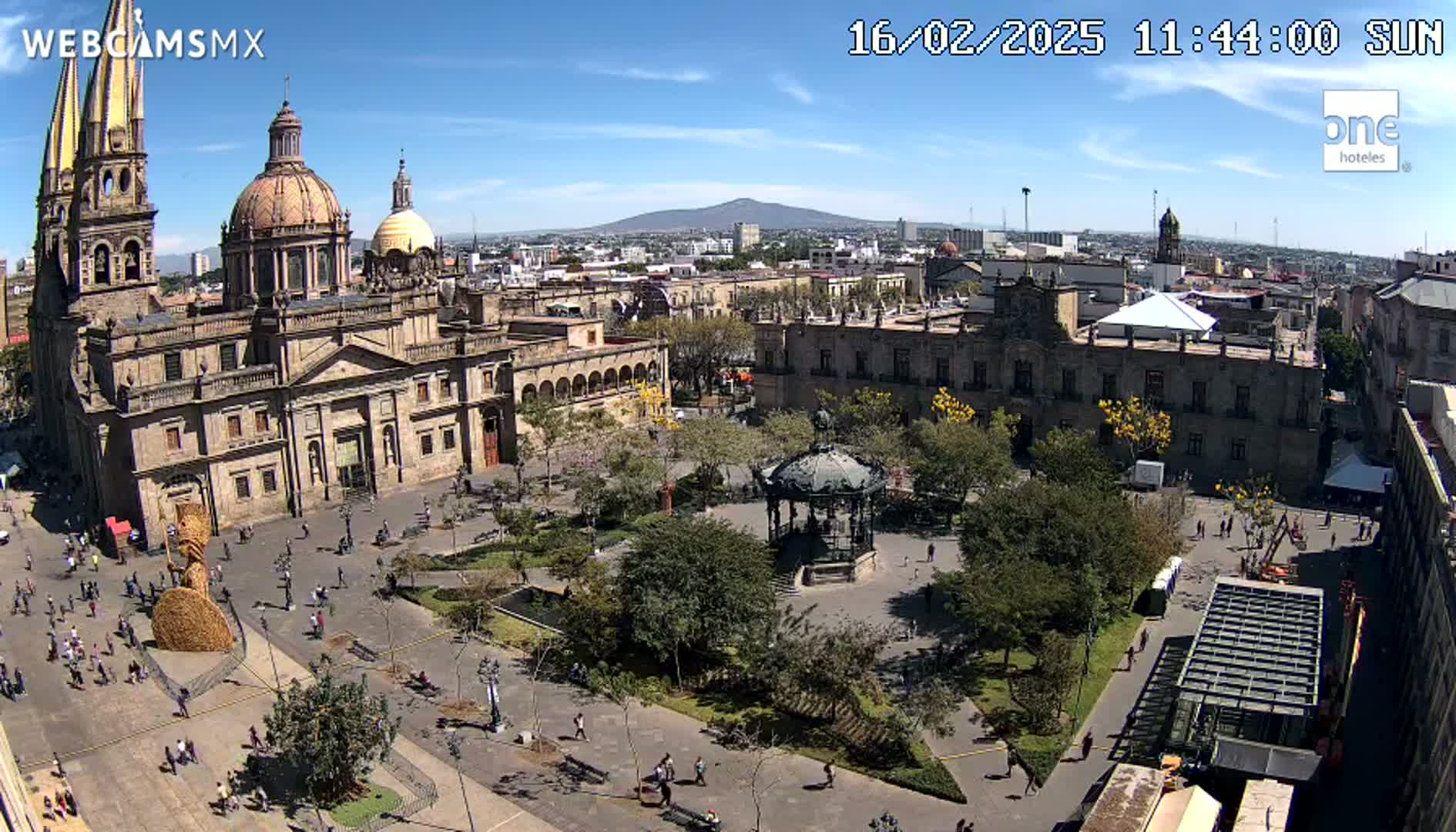 A large cathedral and plaza are seen from above on a sunny day, with a distant volcano visible in the background.