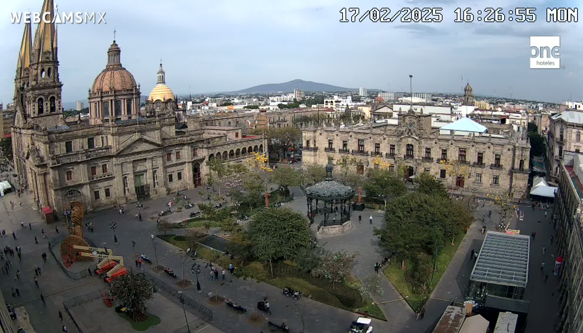 A large plaza with a cathedral and government building, surrounded by trees and people, under a mostly clear sky with a distant mountain visible.