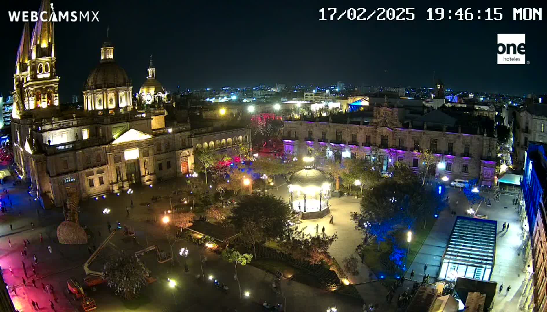 A brightly lit plaza at night, featuring a large cathedral and surrounding buildings, with a gazebo in the center.