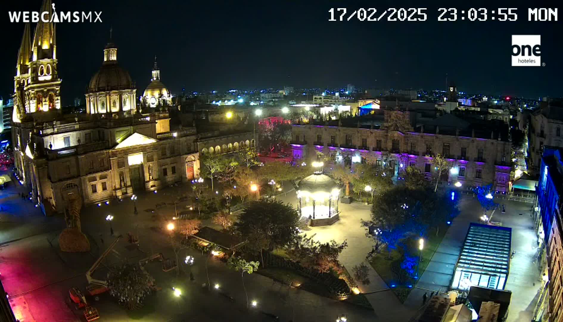 A nighttime, aerial view of a city square, illuminated with lights, featuring a large cathedral and a gazebo, under a clear sky.