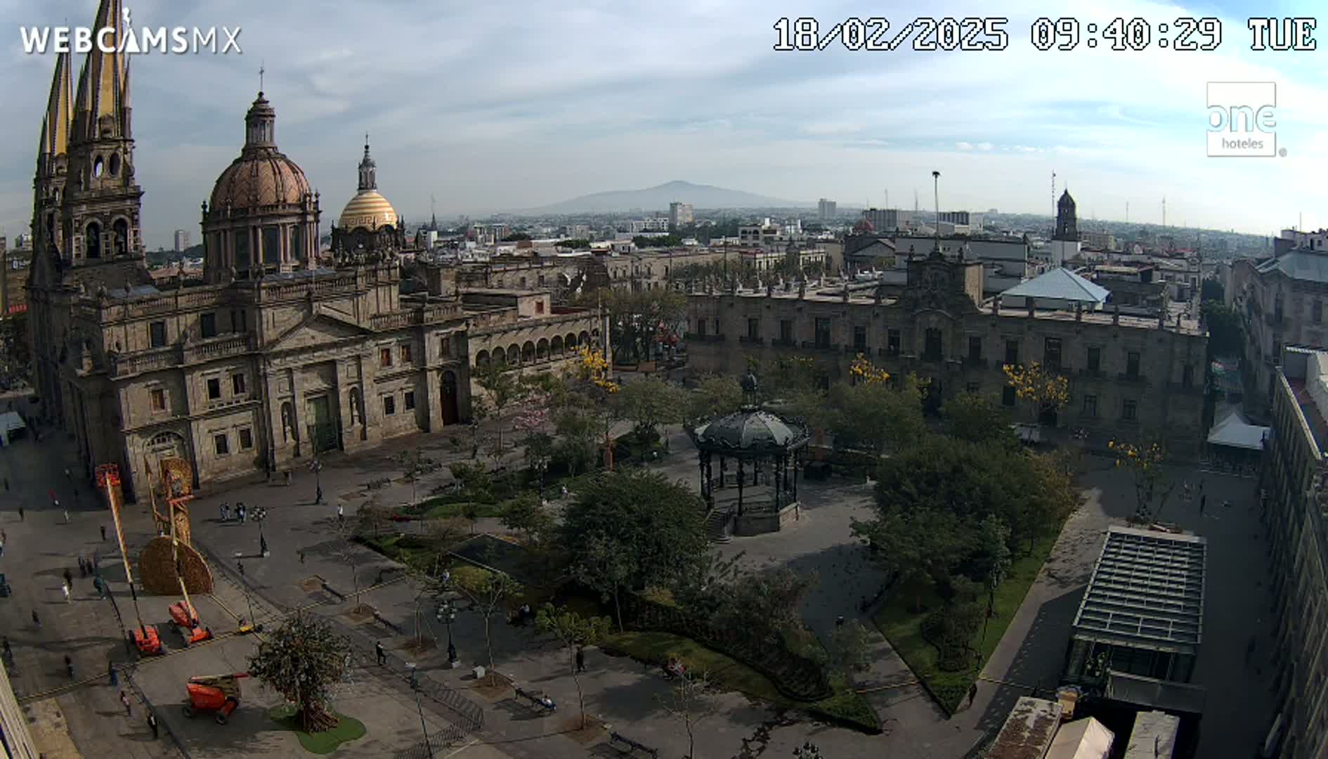 A large cathedral and surrounding plaza are visible on a partly cloudy day, with a distant mountain range in the background.