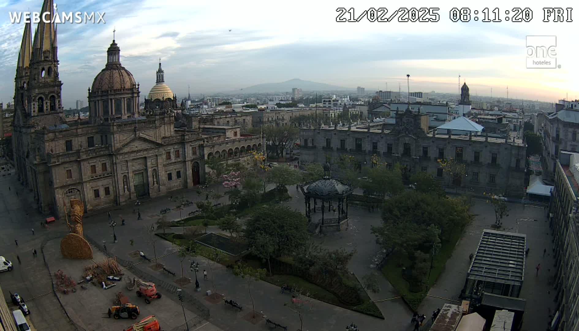 A large cathedral and plaza are visible in a city under a mostly cloudy sky, with a distant mountain range in the background.