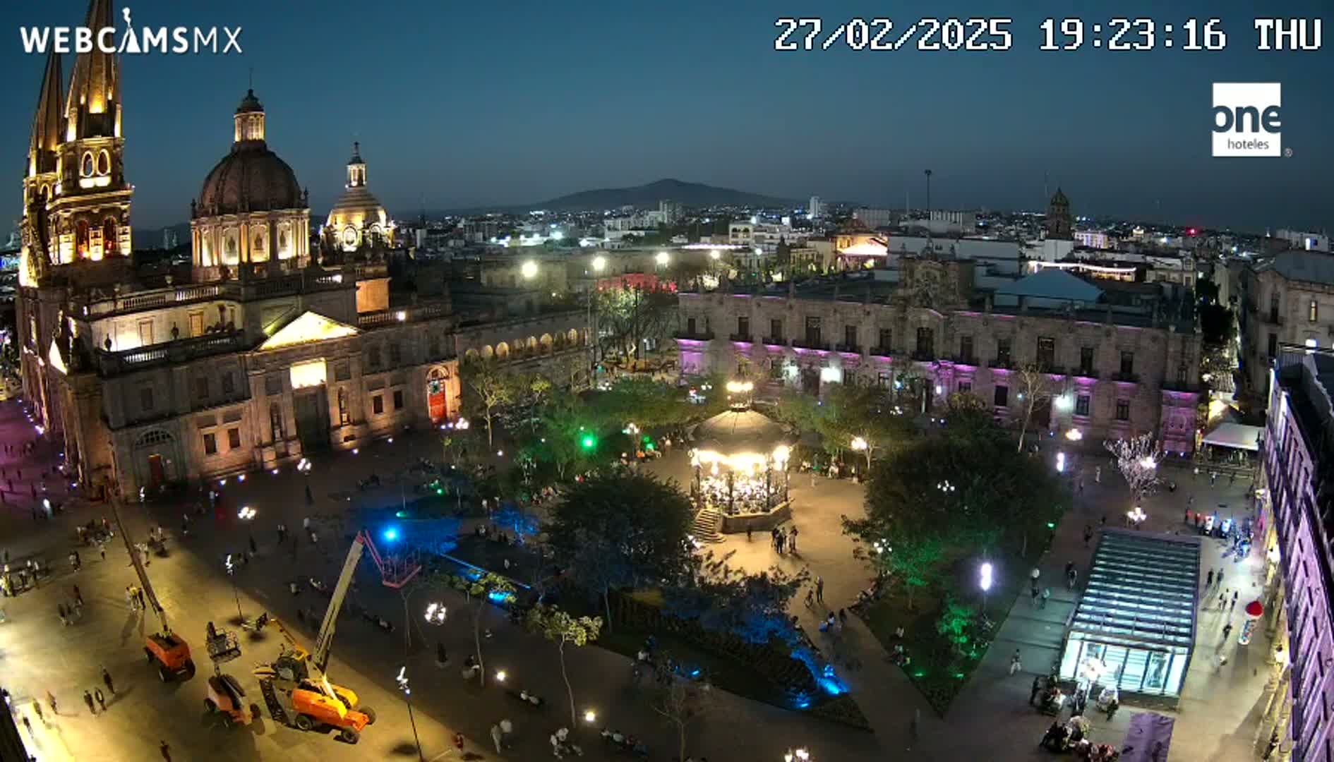A large, illuminated plaza at night, featuring a cathedral, a gazebo, and many people, under a clear, dark sky.