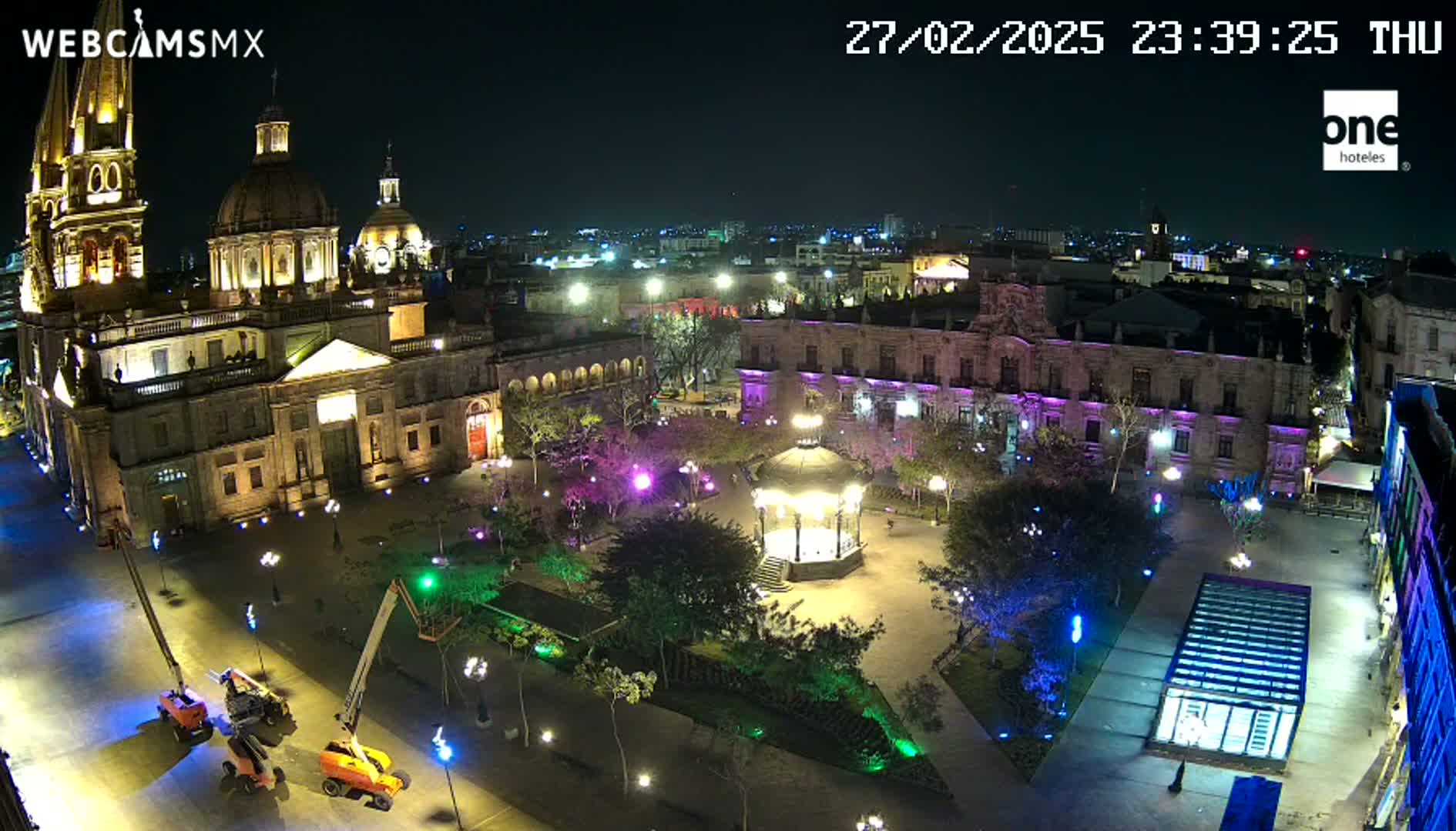 A nighttime aerial view of a plaza with a large cathedral, surrounding buildings, and a gazebo, illuminated by artificial lights.
