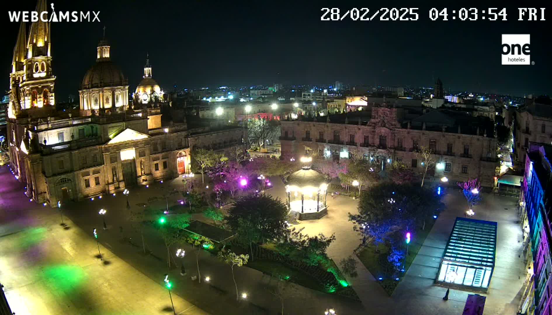 A brightly lit plaza at night, centered around a gazebo, with a large cathedral to one side and other buildings surrounding it under a clear, dark sky.