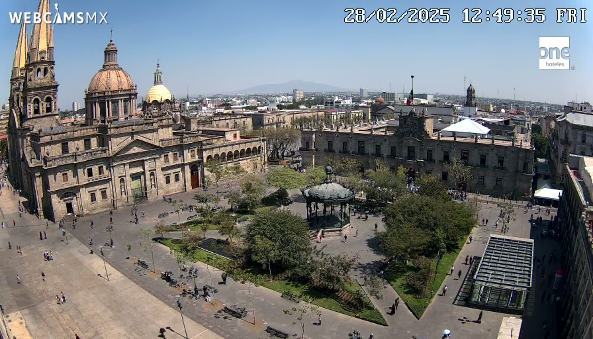 A large plaza with a cathedral and other buildings is seen on a sunny day, with a few people walking around.