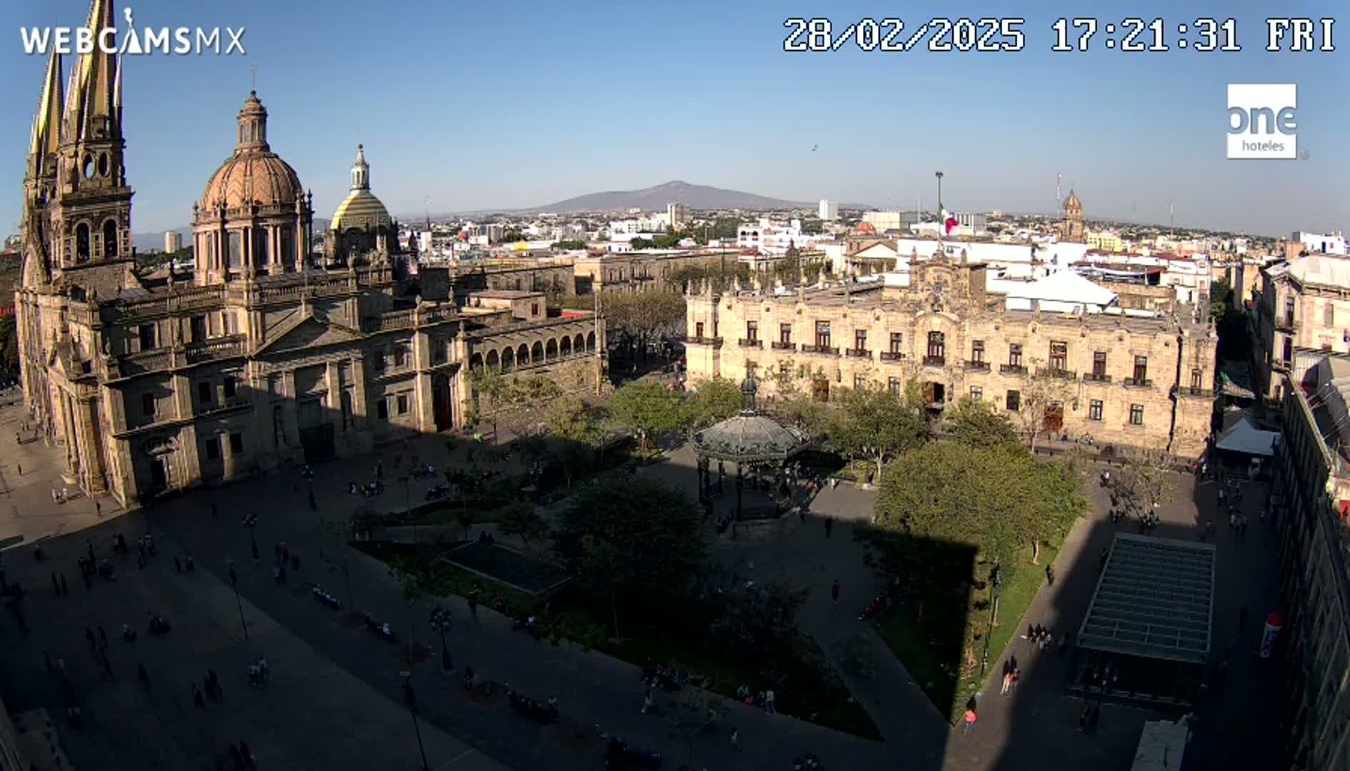 A large plaza with a cathedral and government buildings is shown under sunny conditions, with a distant mountain visible in the background.