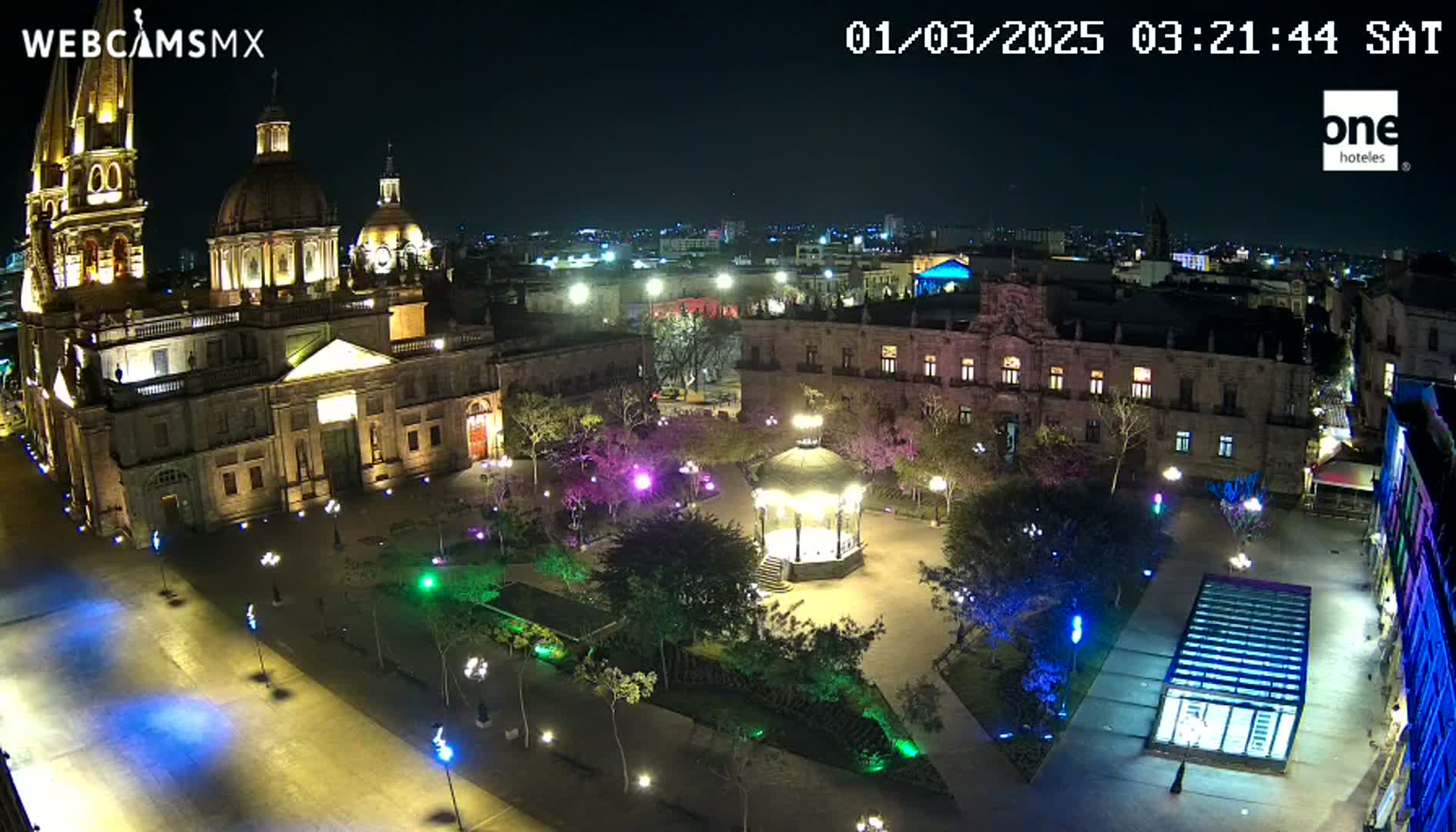 A brightly lit plaza at night, featuring a large cathedral, government building, gazebo, and surrounding trees, under a clear sky.