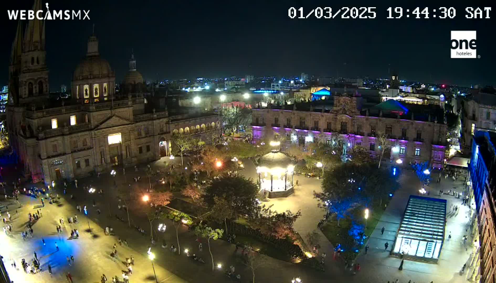 An illuminated plaza at night, featuring a cathedral, surrounding buildings, a gazebo, and many people, under a clear sky.