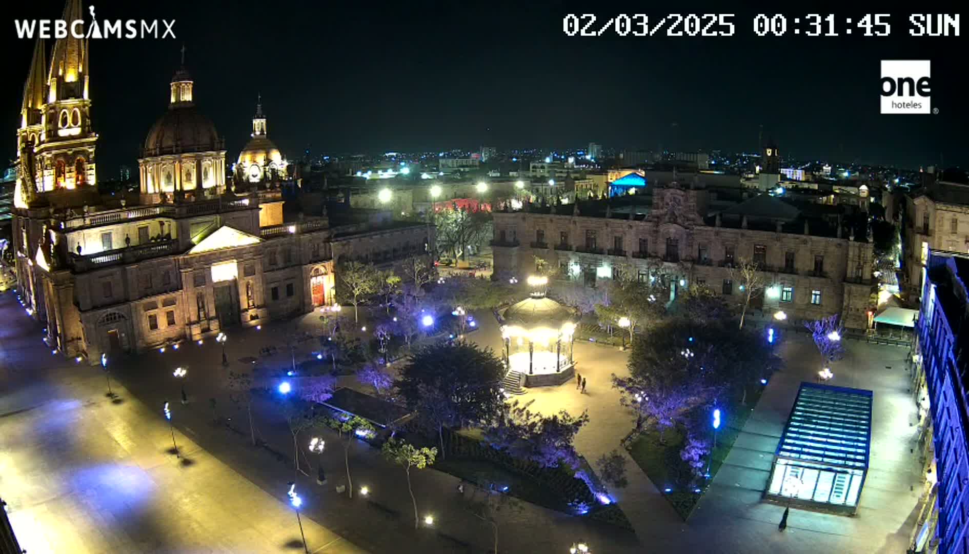 A nighttime aerial view shows a large plaza with a gazebo, surrounded by illuminated buildings, including a cathedral, under a clear sky.