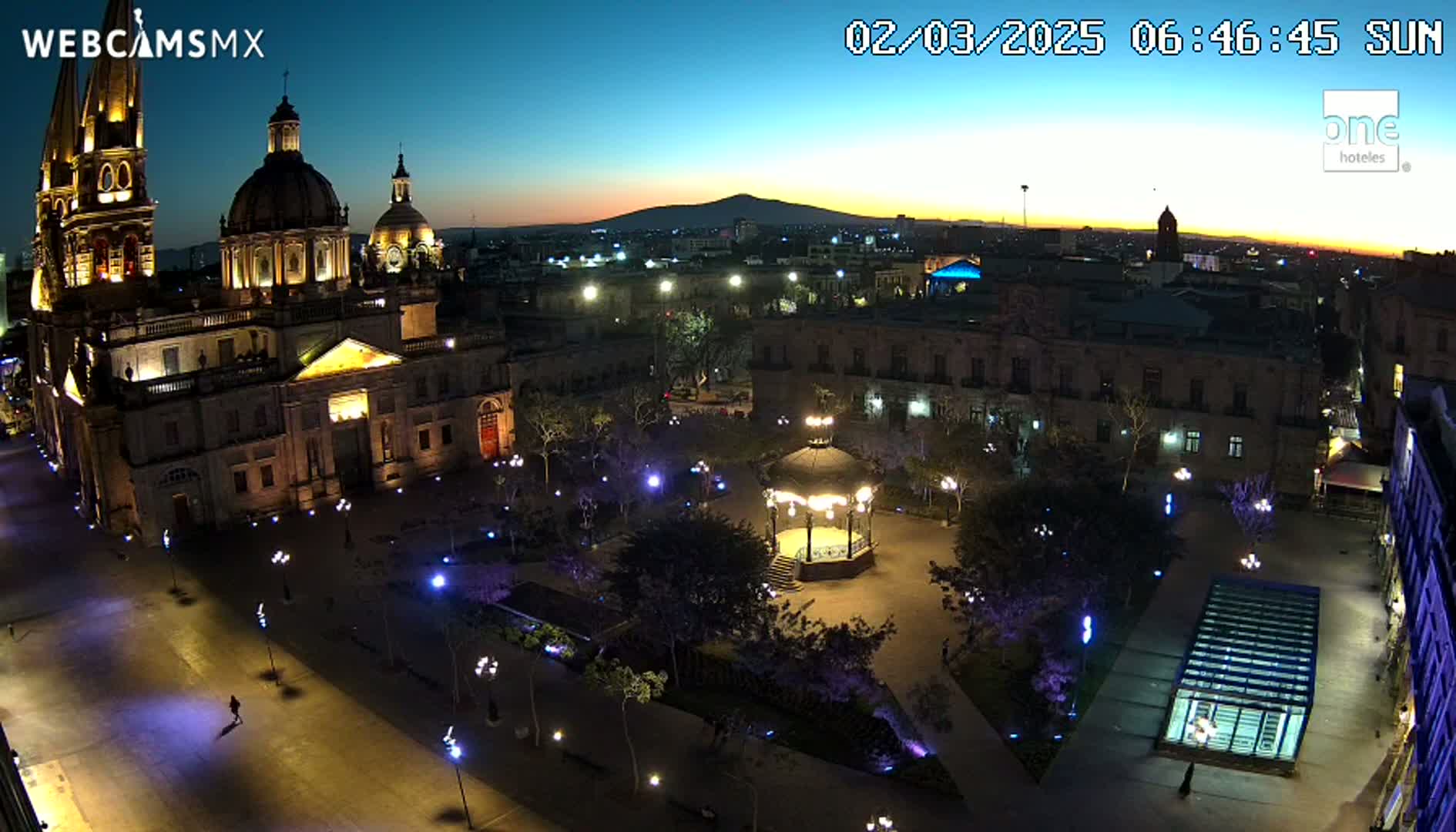An illuminated plaza at twilight, featuring a large cathedral and gazebo, with a mountain range visible in the distance under a clear sky.