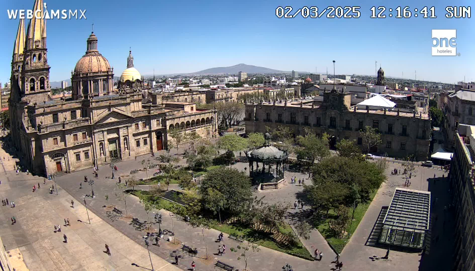 A high-angle view of a large plaza in a city on a sunny day, featuring a cathedral, government buildings, a gazebo, and many trees and people.
