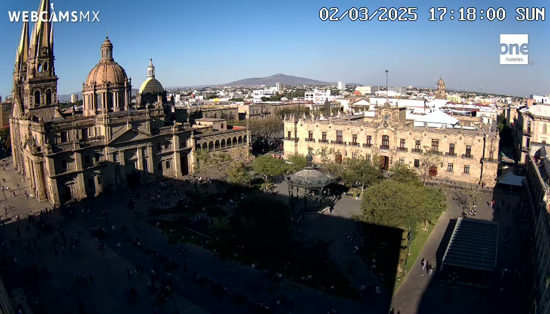 A large cathedral and government building are situated in a sunny city square filled with people, with a distant mountain visible under a clear blue sky.