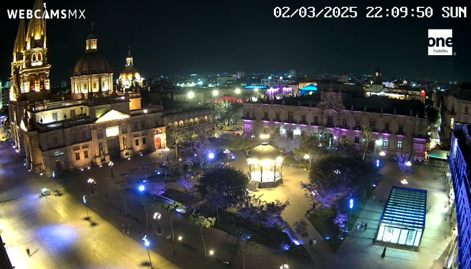A brightly lit plaza at night, featuring a large cathedral, surrounding buildings, a gazebo, and sparsely populated walkways under a clear night sky.