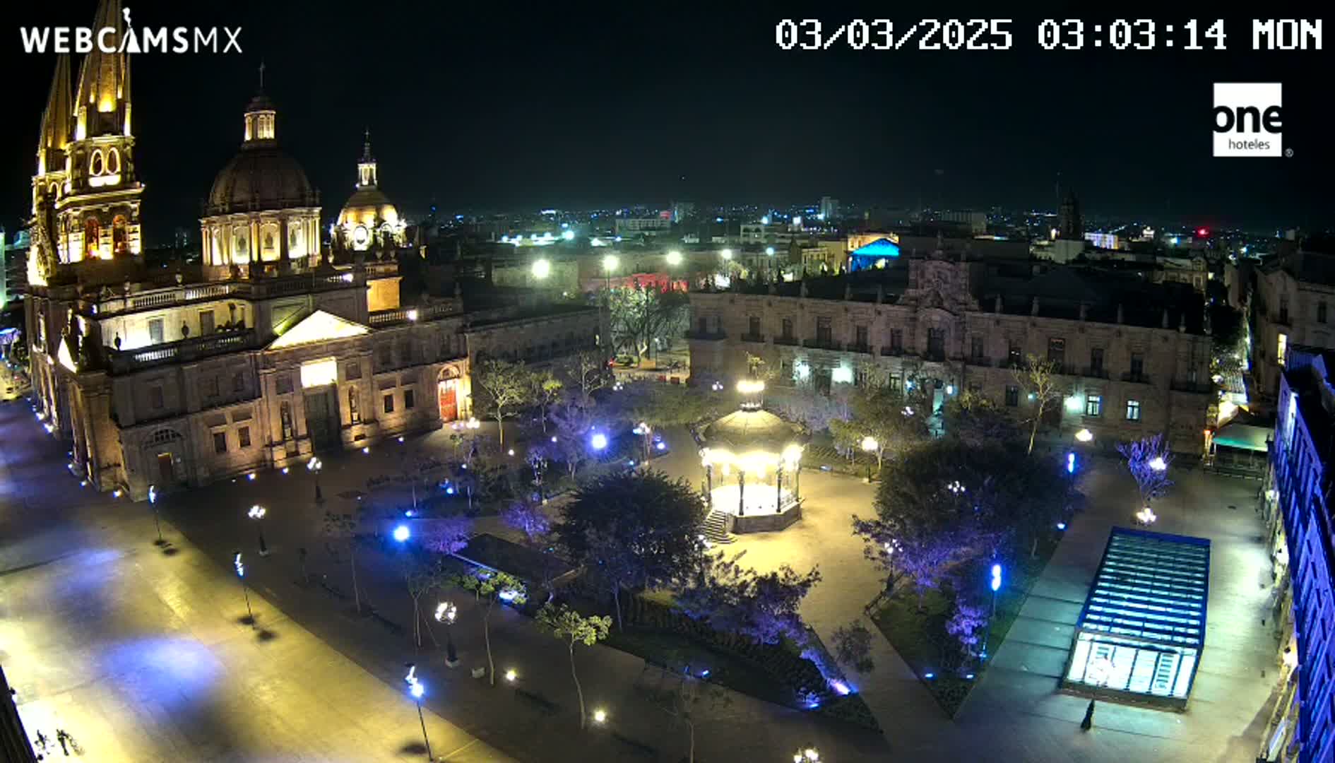 A brightly lit plaza at night, centered around a gazebo, with a large cathedral and surrounding buildings visible, under a clear night sky.