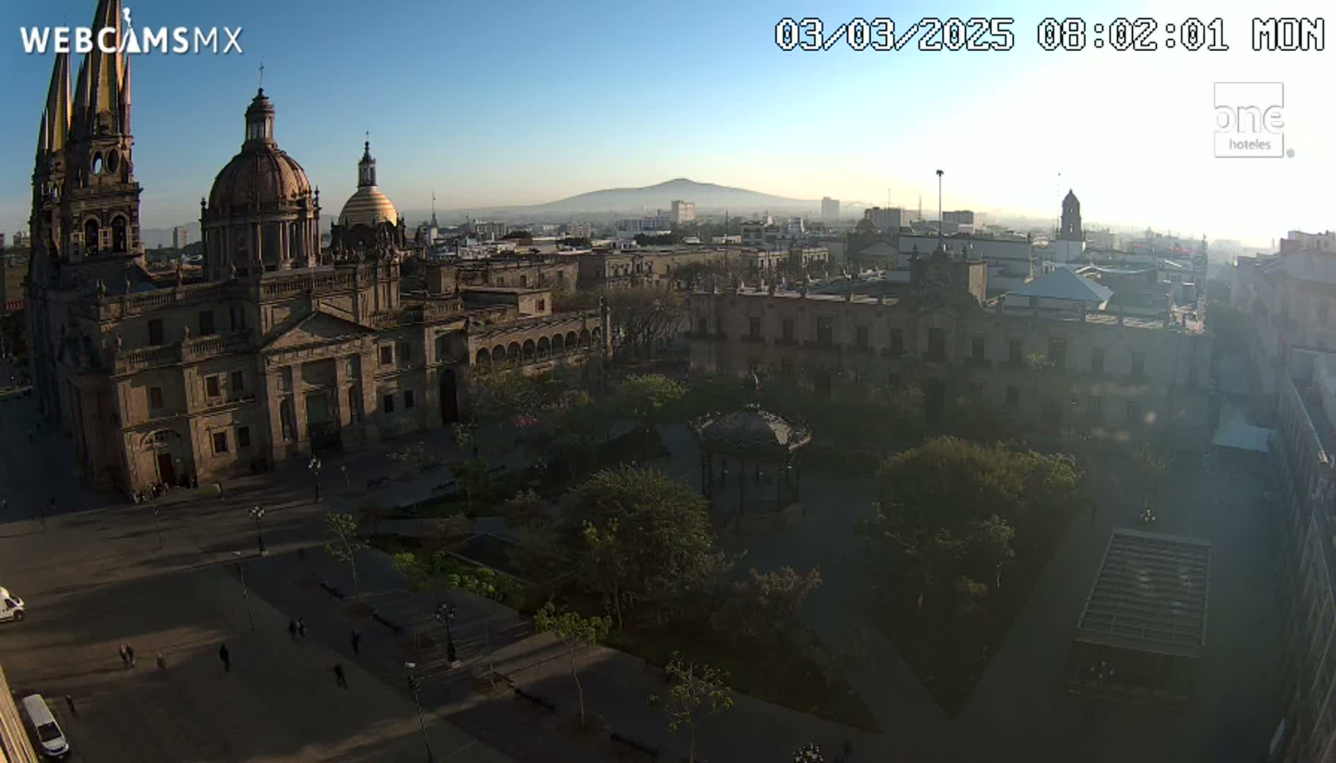 A large cathedral and surrounding plaza are visible in the early morning, with a hazy mountain in the background under a clear blue sky.