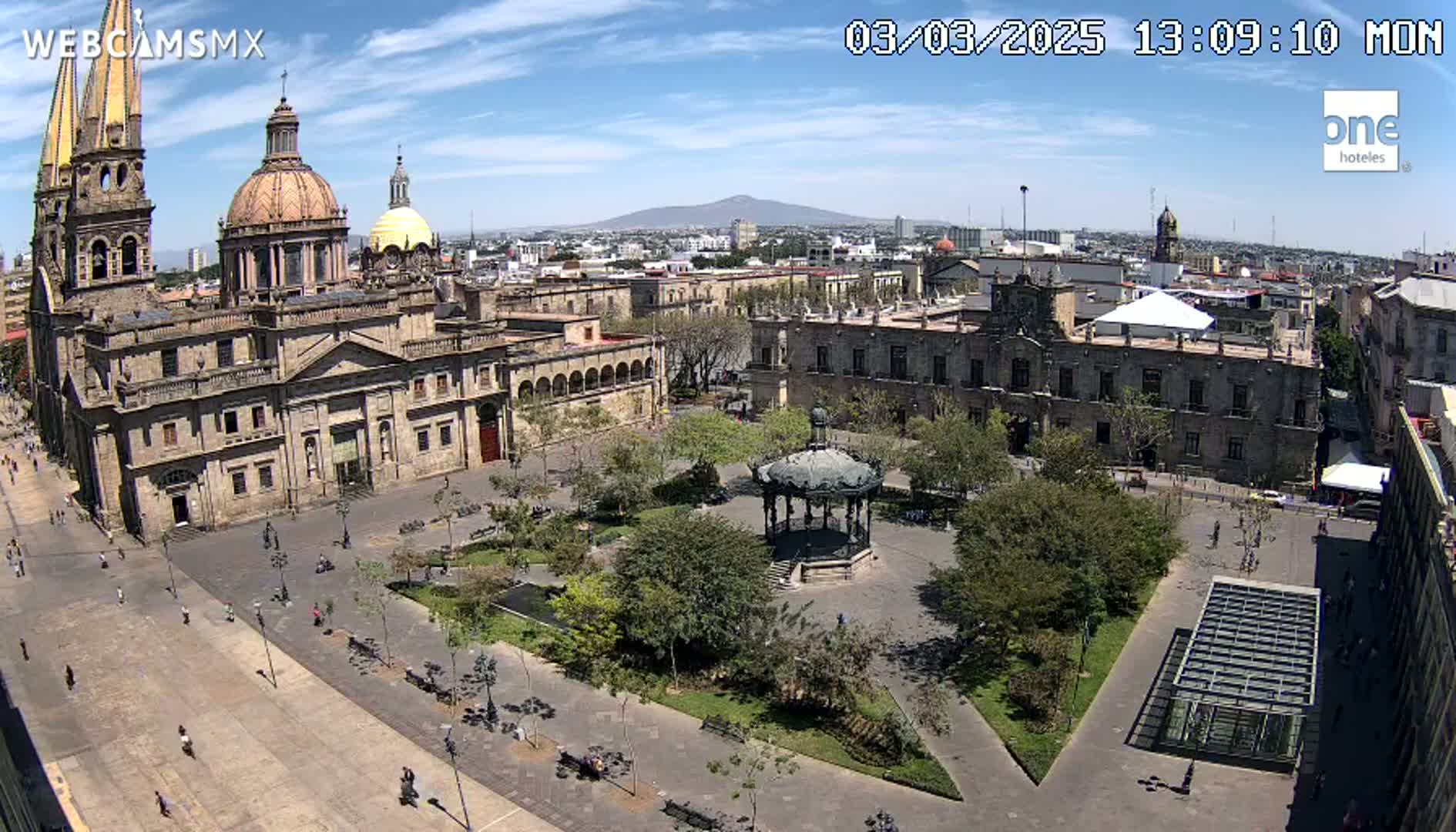 A large plaza with a gazebo, surrounded by historic buildings, including a cathedral, under a mostly sunny sky with a distant volcano visible in the background.