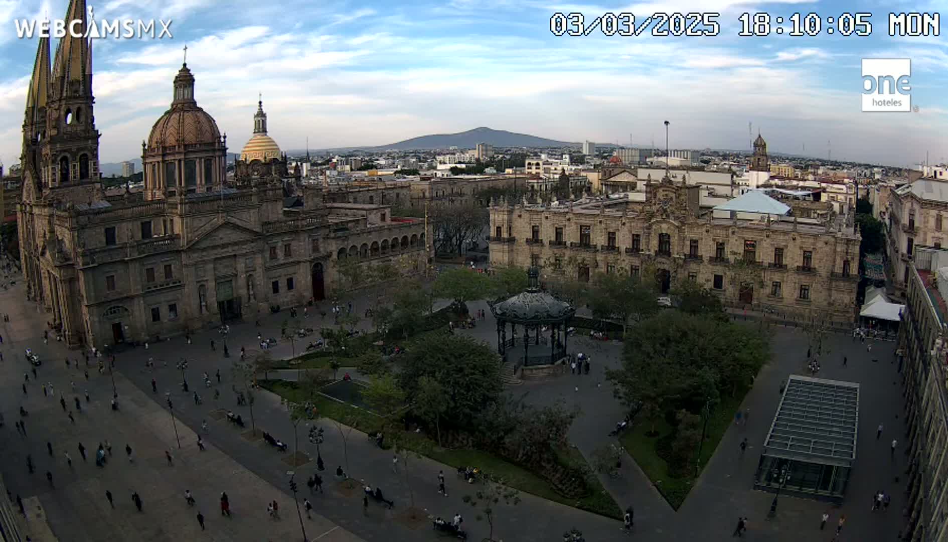 An aerial view of a large plaza in a city, under a mostly clear sky, shows a cathedral and government buildings surrounding a central park with a gazebo and many people walking around.
