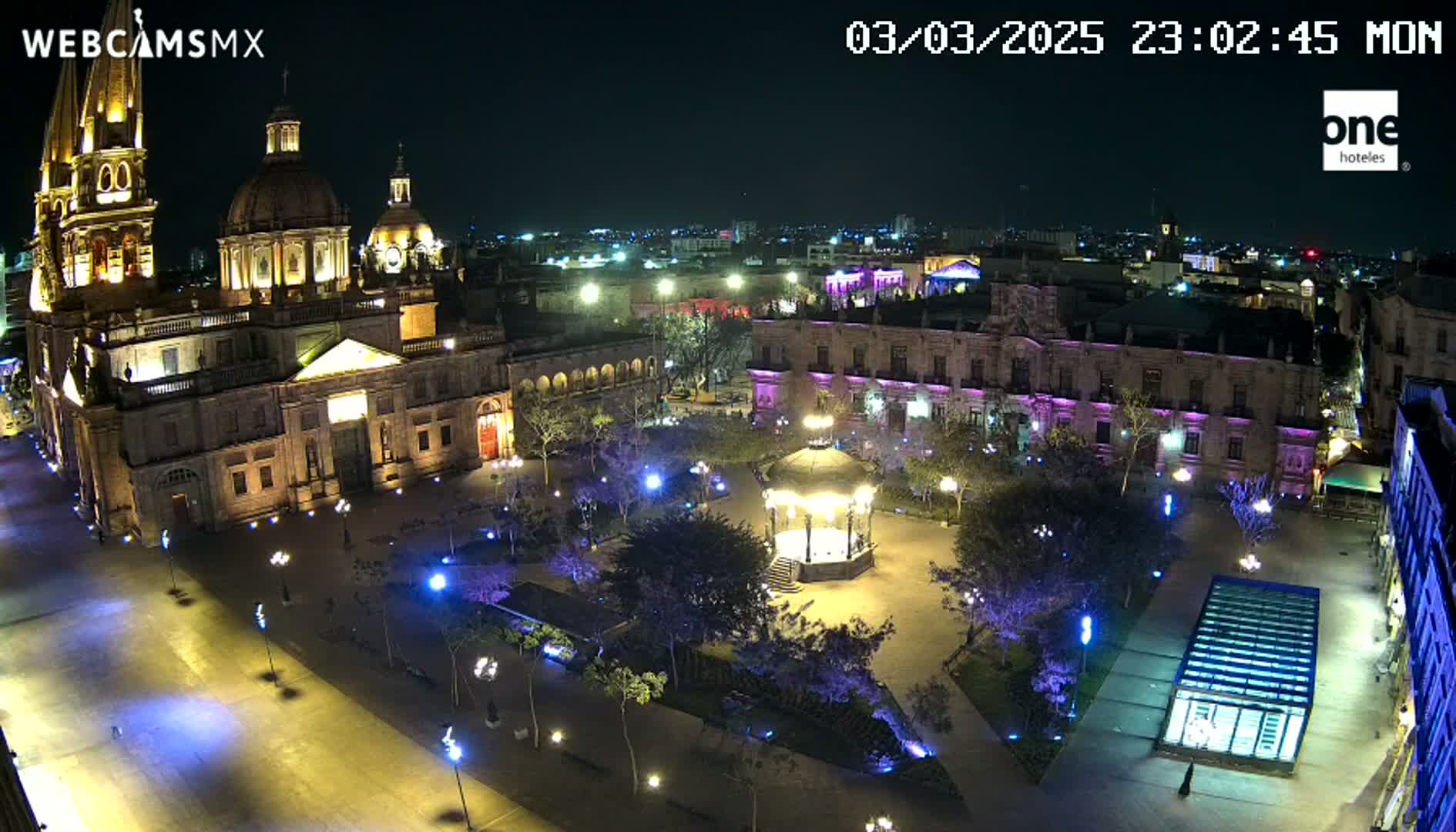 A brightly lit plaza at night, featuring a large cathedral, surrounding buildings, a gazebo, and many trees.