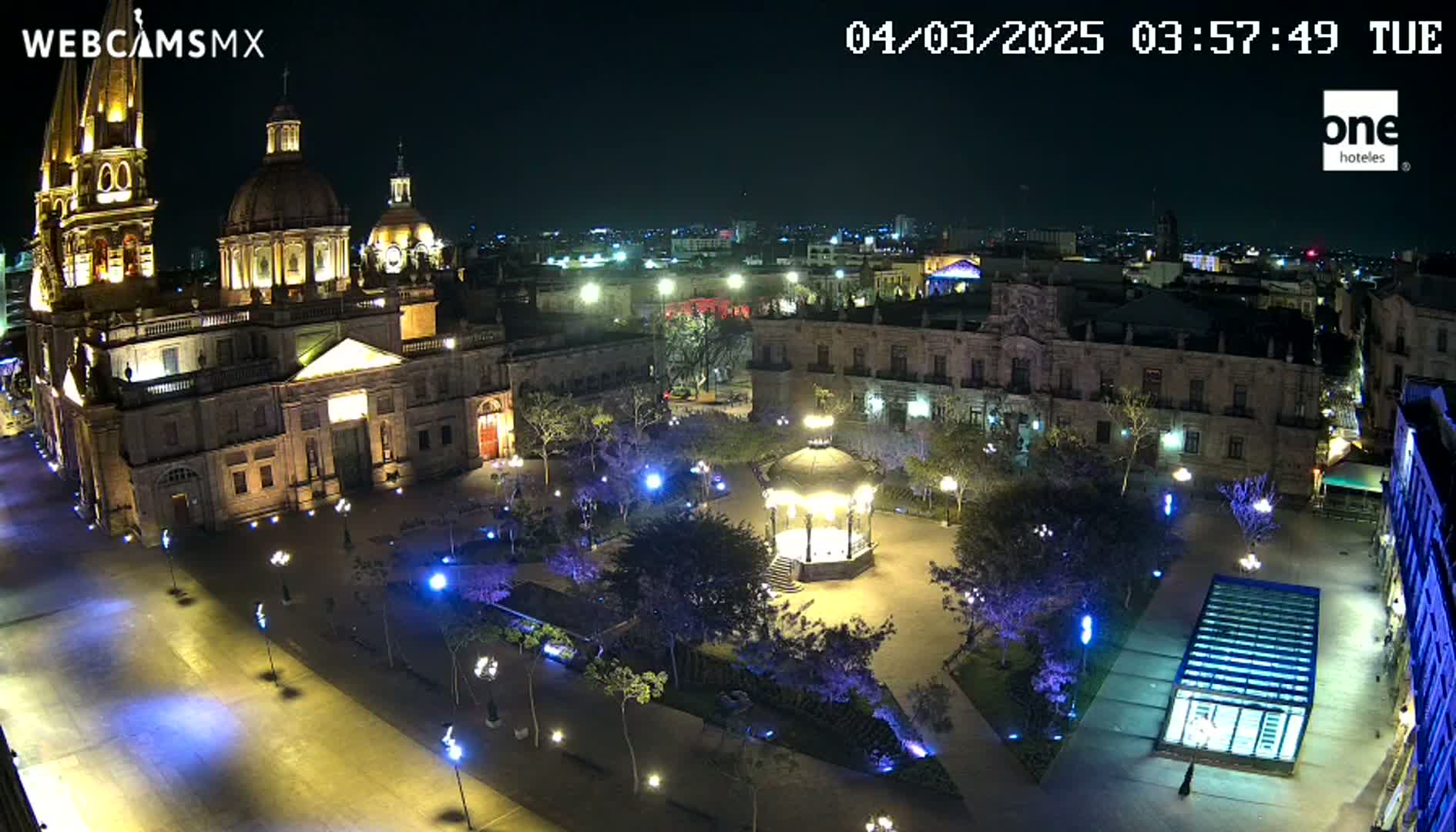 A brightly lit plaza at night features a large cathedral, surrounding buildings, a gazebo, and numerous trees and lights.