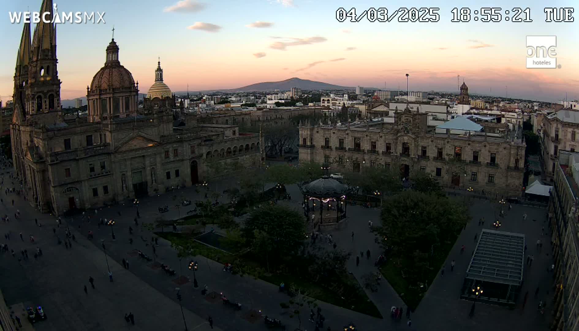 An aerial view of a city square at dusk, showing a large cathedral, government buildings, a gazebo, and people walking around, with a mountain visible in the distance under a pinkish-purple sky.