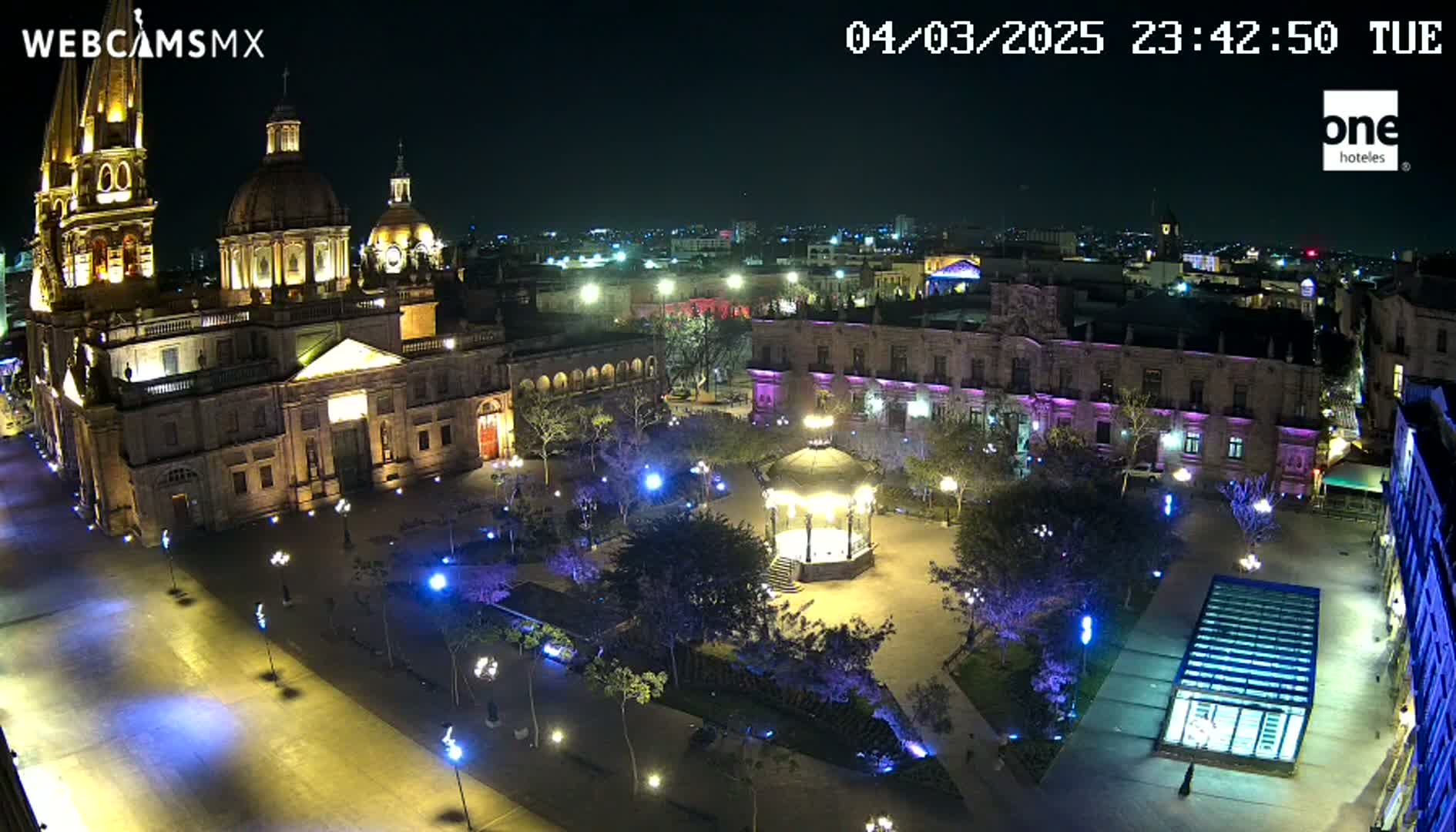 A brightly lit plaza at night, featuring a large cathedral and surrounding buildings, with a gazebo in the center of the square.