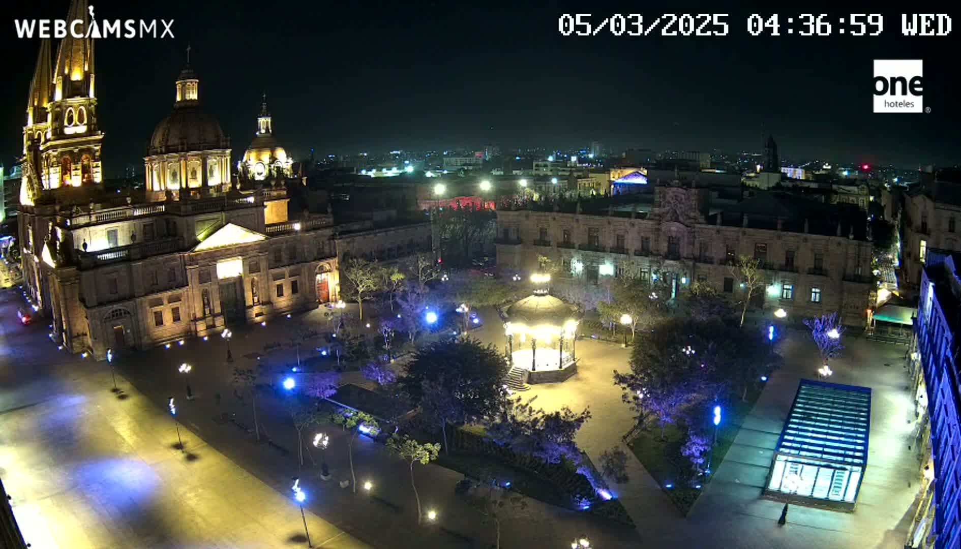 A brightly lit plaza at night features a large cathedral, surrounding buildings, and a gazebo in the center, under a clear, dark sky.