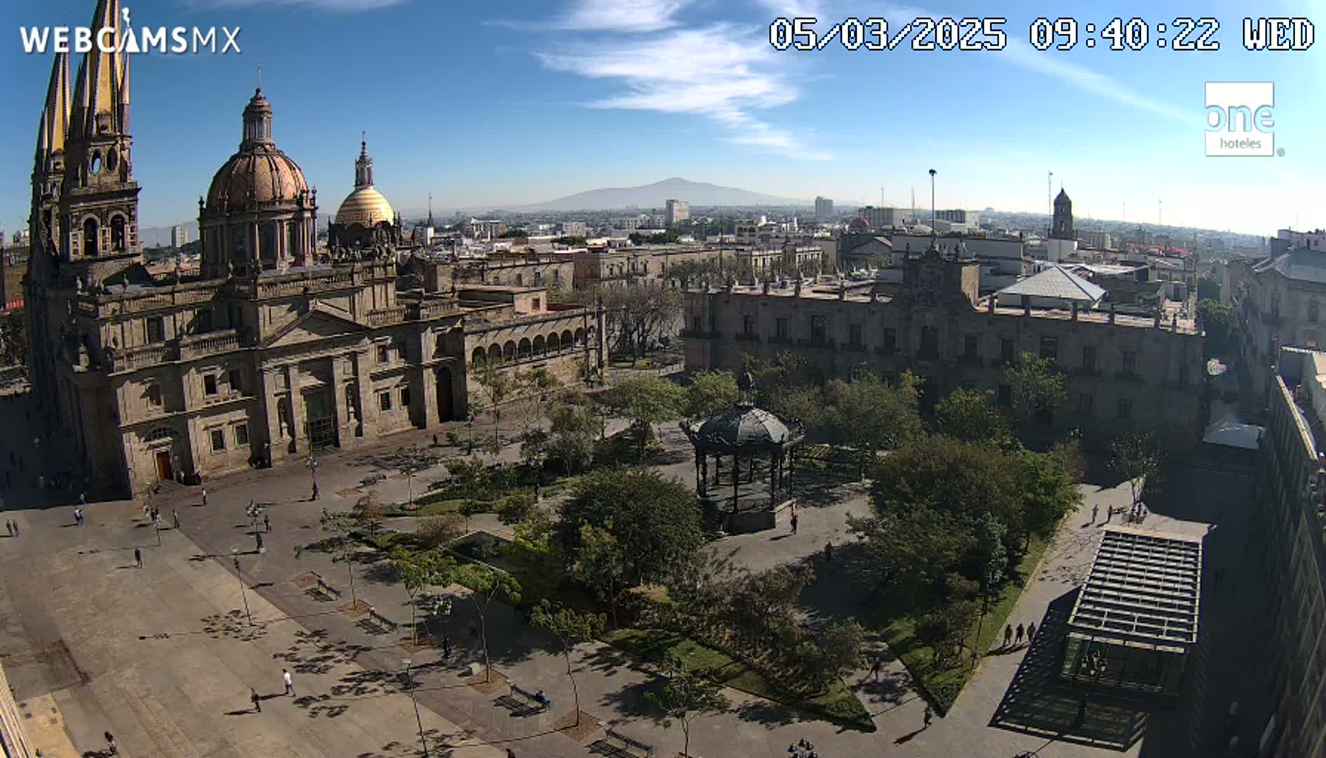 A large cathedral and plaza, with a gazebo and surrounding trees, are seen under a clear, sunny sky, with a distant mountain visible on the horizon.