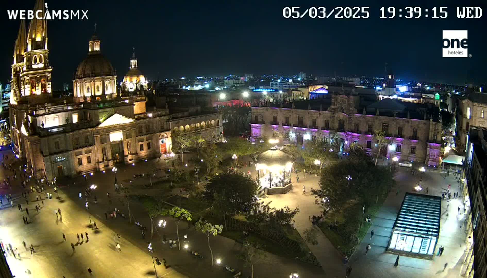 A brightly lit plaza at night, featuring a large cathedral, other buildings, a gazebo, and many people walking around.